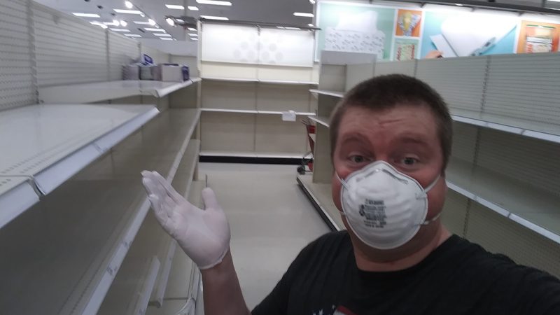 A man wearing an N95 mask and gloves stands in a aisle between empty shelves.