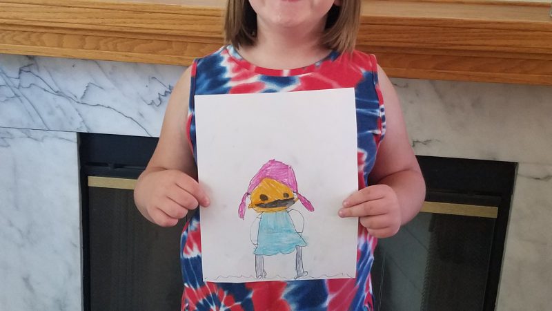 A young girl stands in front of a fireplace holding a drawing of a child wearing a mask.