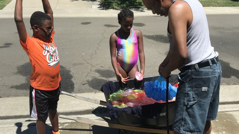 A man and two children stand around a wheelbarrow filled with different colored water balloons.