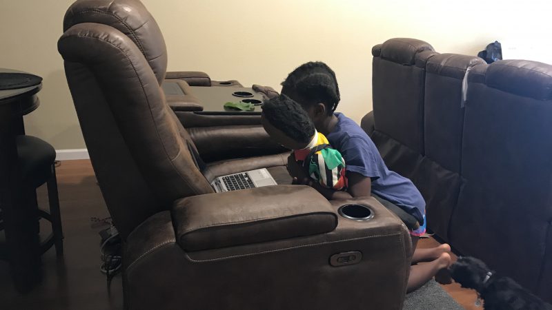 A young girl with a doll kneels in front of an open laptop on a brown reclining chair.