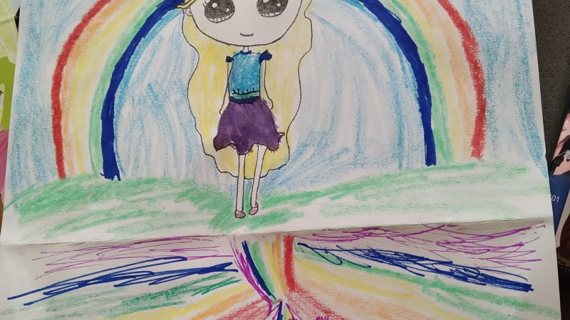 A children's drawing of a girl with long blonde hair standing in front of a rainbow, with two unicorns beneath her.