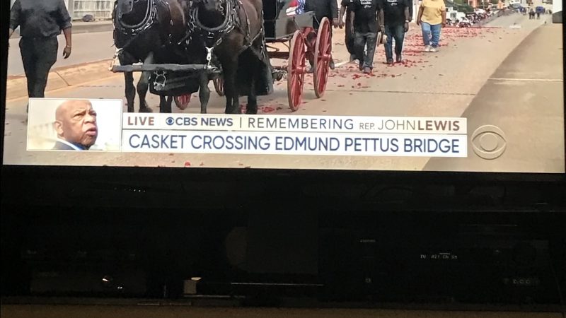 Photograph of television screen showing a funeral procession led by a carriage pulled by two horses crossing over a bridge.