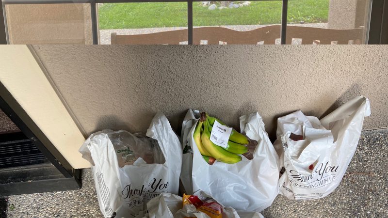 The top half of the photo shows a delivery man in front of a delivery truck as seen through a window. The bottom half shows five plastic bags full of groceries on the ground.