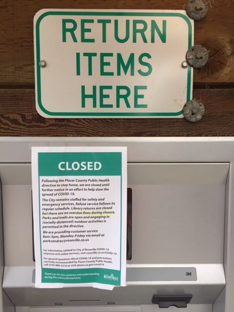 A green and white sign on a library bookdrop notes that the library is closed.