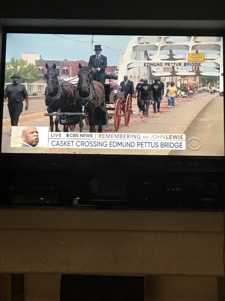 Photograph of television screen showing a funeral procession led by a carriage pulled by two horses crossing over a bridge.