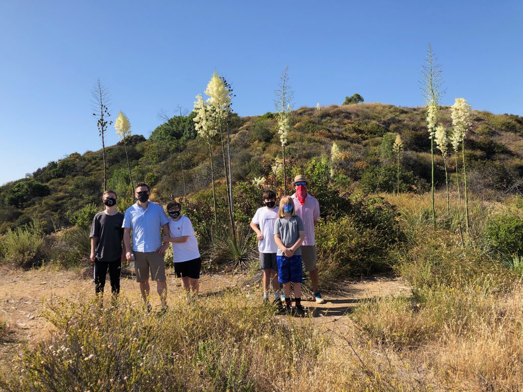 Two adults and four children, all wearing t-shirts, shorts and face masks, stand on a trail in front of a small hill.