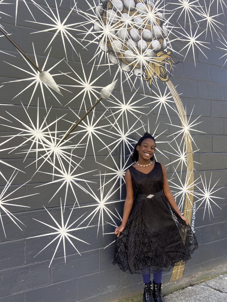 A smiling young girl wearing a black lace dress stands in front of a black wall with stars painted on it.