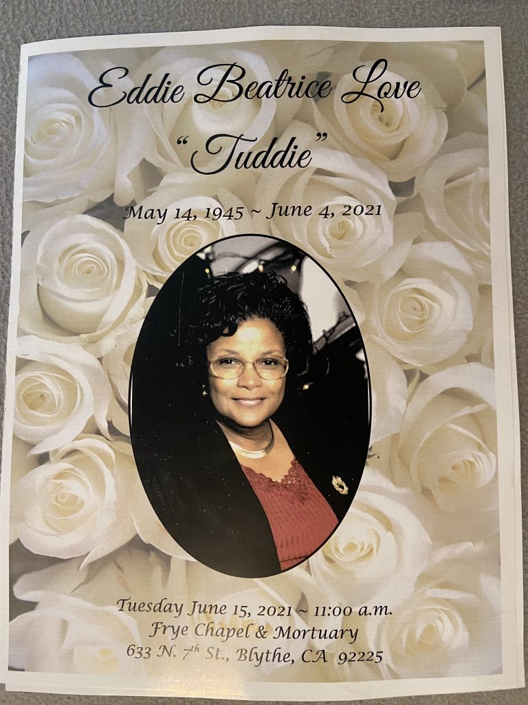 A pamphlet with a photograph of a woman with dark hair and glasses, smiling, against a background of white roses. The pamphlet reads "Eddie Beatrice Love 'Tuddie' May 14, 1945-June 4, 2021."